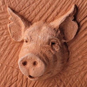 Pode incluir: Escultura em relevo castanho-avermelhada detalhada de uma cabeça de porco. O focinho, os olhos e as orelhas do porco são claramente definidos, com detalhes texturizados no rosto. O fundo tem um padrão diagonal esculpido.
