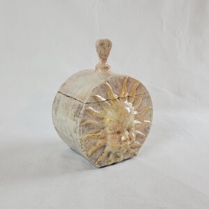 Puede incluir: Caja de cerámica con forma de sol con tapa y asa. La caja es principalmente beige con detalles amarillos en la cara y los rayos del sol. La caja mide aproximadamente 15 cm de diámetro y 13 cm de alto.