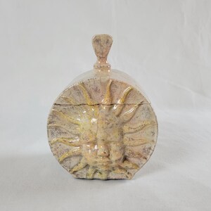 Op de afbeelding: Een ronde keramische doos met deksel in een beige en gele craquelé glazuurafwerking. De voorzijde toont een verhoogd reliëf van een gezicht van de zon met stralen. Een kleine, verticale, schoppenvormige handgreep zit op het gebogen deksel.