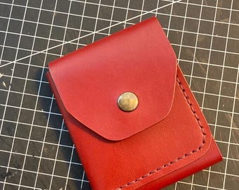 Op de afbeelding: Een kleine, rode leren portemonnee met een klepsluiting die is vastgezet met een messingkleurige drukknop. De randen zijn netjes gestikt met een contrasterende draad. De portemonnee is waarschijnlijk ontworpen om kaarten of kleine items in te bewaren.