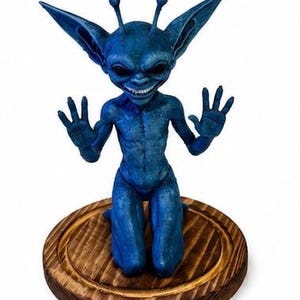 May include: A blue creature figurine with large pointed ears, two antennae, and black eyes, kneeling with hands raised. The figure has a textured surface and a wide grin showing teeth, mounted on a circular, dark-stained wooden base.
