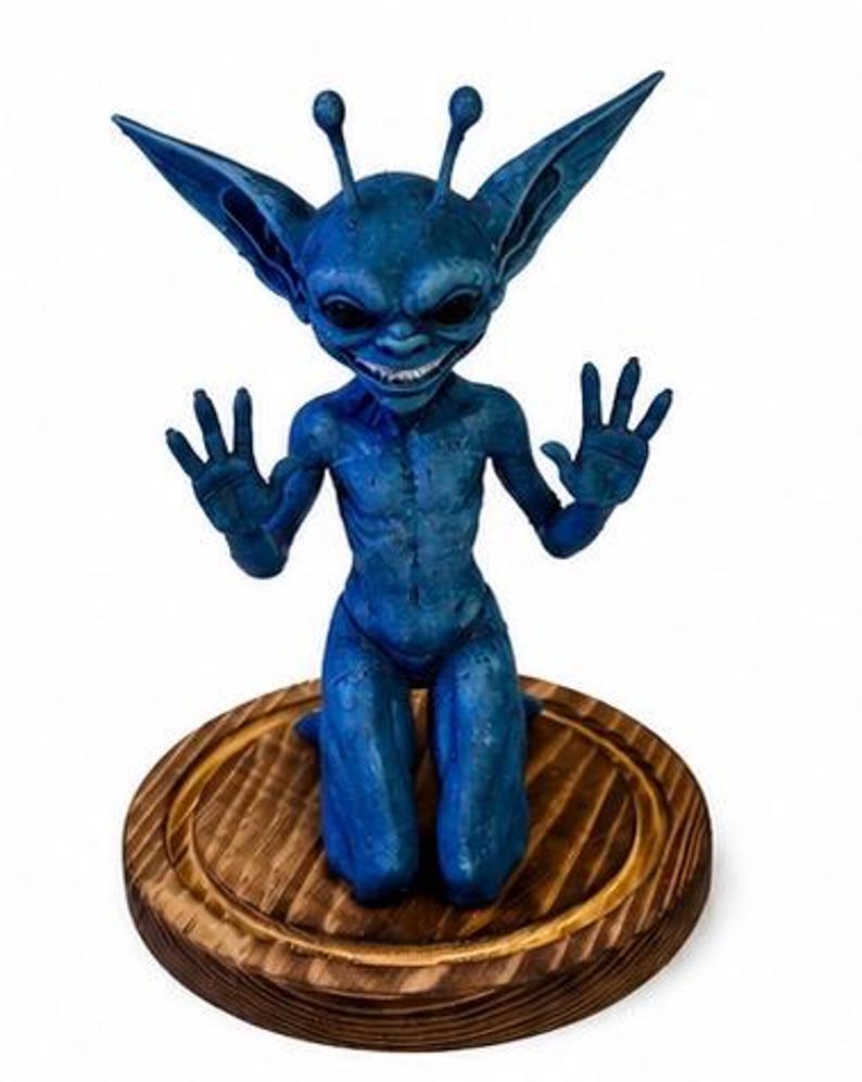 May include: A blue creature figurine with large pointed ears, two antennae, and black eyes, kneeling with hands raised. The figure has a textured surface and a wide grin showing teeth, mounted on a circular, dark-stained wooden base.