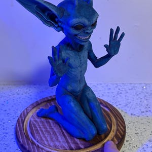 May include: A blue, goblin-like creature figurine with large, pointed ears and antennae, kneeling on a circular wooden base. The figure has a wide, toothy grin, dark eyes, and raised hands with fingers spread, rendered in a textured, matte blue finish.