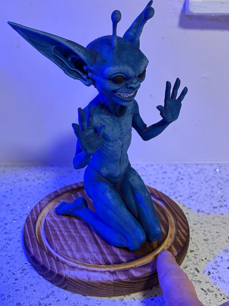 May include: A blue, goblin-like creature figurine with large, pointed ears and antennae, kneeling on a circular wooden base. The figure has a wide, toothy grin, dark eyes, and raised hands with fingers spread, rendered in a textured, matte blue finish.