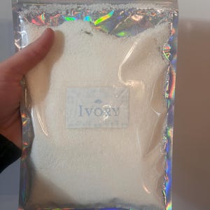 May include: A clear, iridescent bag filled with white granules. The bag has a silver zipper and a label that reads "Ivory" in a light blue font. The bag is held by a person.