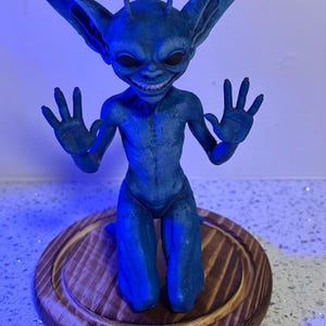 May include: A blue creature figurine with large pointed ears, two antennae, and a wide grin showing teeth. The creature is in a kneeling position with both hands raised, palms facing forward. It is mounted on a circular, stained wooden base.
