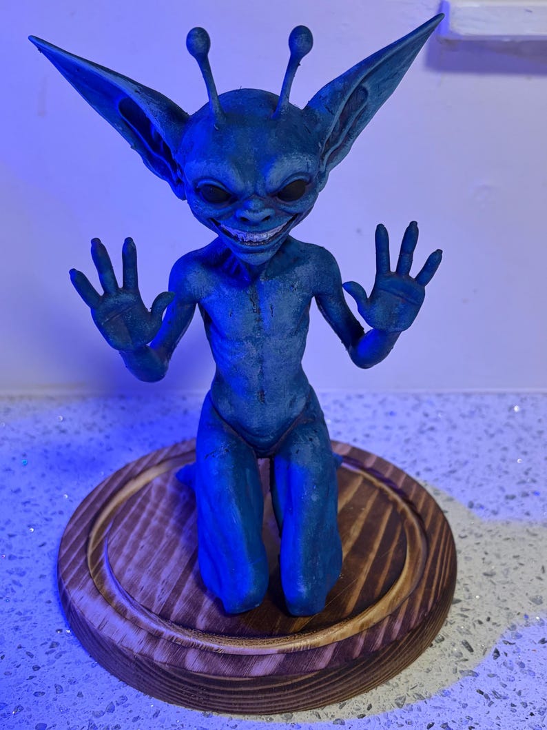 May include: A blue creature figurine with large pointed ears, two antennae, and a wide grin showing teeth. The creature is in a kneeling position with both hands raised, palms facing forward. It is mounted on a circular, stained wooden base.