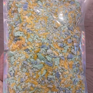 May include: A clear plastic bag filled with colorful, irregularly shaped pieces. The pieces are primarily yellow, blue, and green, with a few white fragments. The bag has a holographic sheen.