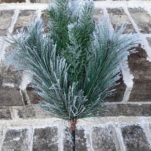 May include: A green and white artificial pine branch with a frosted appearance. The branch is on a black metal stem, set against a brick wall. The pine needles are covered in a white, icy-looking substance.