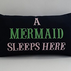 May include: A black pillow with the text "A Mermaid Sleeps Here" embroidered in pink and green.