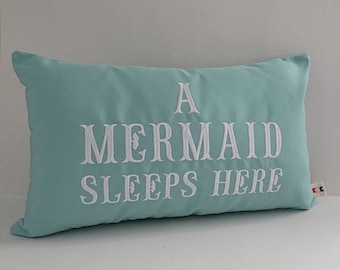 May include: A turquoise blue pillow with white embroidered text that reads "A Mermaid Sleeps Here".