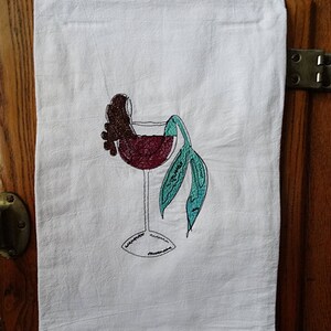 May include: A white kitchen towel with an embroidered design of a wine glass with a mermaid tail and a bunch of grapes. The mermaid tail is turquoise and the grapes are brown.