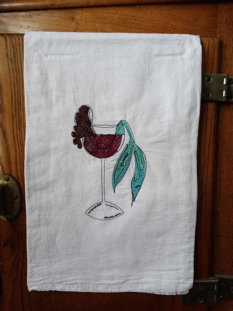 May include: A white kitchen towel with an embroidered design of a wine glass with a mermaid tail and a bunch of grapes. The mermaid tail is turquoise and the grapes are brown.