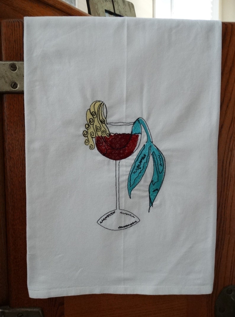 May include: A white kitchen towel with an embroidered design of a mermaid in a wine glass. The mermaid has blonde hair and a turquoise tail. The wine glass is filled with red wine.