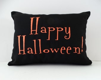 Può includere: Un cuscino rettangolare nero con la scritta Happy Halloween! ricamata in filo arancione brillante. Il testo è centrato sulla parte anteriore del cuscino, con la parola Happy sopra Halloween e un punto esclamativo alla fine della frase.