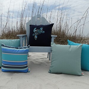 May include: Four decorative throw pillows on a white sandy beach. One pillow has a blue and green striped pattern, another has a solid teal color, and the third has a solid light blue color. The fourth pillow is navy blue with a blue and white sea turtle design.