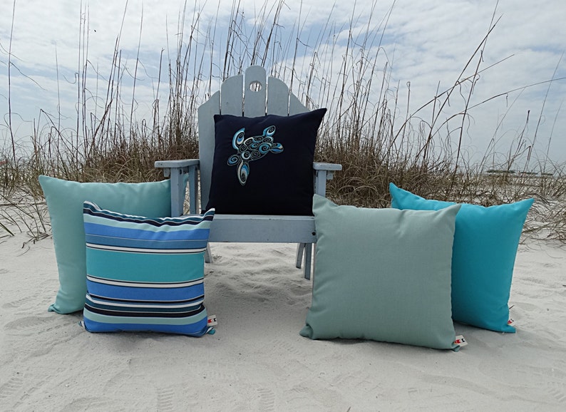 May include: Four decorative throw pillows on a white sandy beach. One pillow has a blue and green striped pattern, another has a solid teal color, and the third has a solid light blue color. The fourth pillow is navy blue with a blue and white sea turtle design.