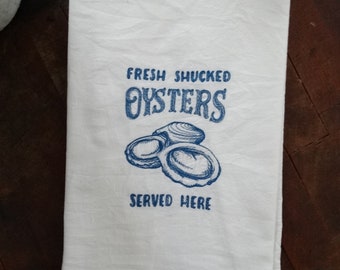 May include: White kitchen towel with blue text and a drawing of oysters. The text reads "Fresh Shucked Oysters Served Here."