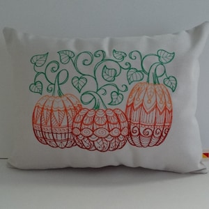 K&ouml;nnte beinhalten: Ein wei&szlig;es Kissen mit einem gestickten Design aus drei orangefarbenen K&uuml;rbissen mit gr&uuml;nen Ranken und Bl&auml;ttern.