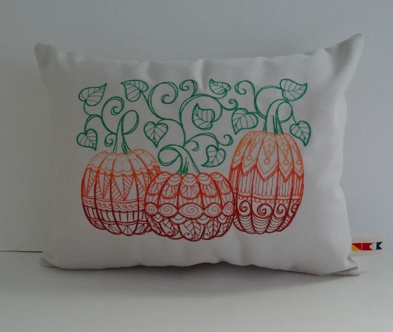 K&ouml;nnte beinhalten: Ein wei&szlig;es Kissen mit einem gestickten Design aus drei orangefarbenen K&uuml;rbissen mit gr&uuml;nen Ranken und Bl&auml;ttern.