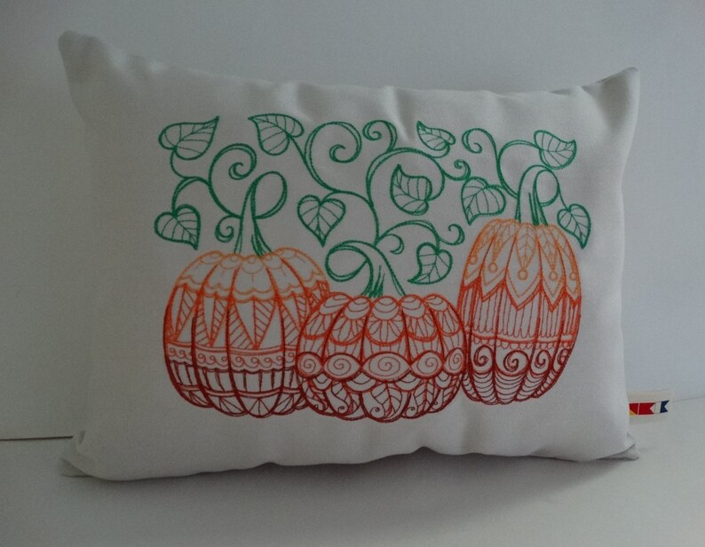 K&ouml;nnte beinhalten: Ein wei&szlig;es Kissen mit einem gestickten Design aus drei orangefarbenen und roten K&uuml;rbissen mit gr&uuml;nen Ranken und Bl&auml;ttern.