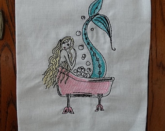 May include: A white linen tea towel with an embroidered design of a mermaid in a pink bathtub with bubbles. The mermaid has long blonde hair and a turquoise tail.