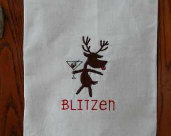 May include: White kitchen towel with a brown embroidered reindeer holding a martini glass with an olive. The reindeer has a red tongue sticking out. The text "Blitzen" is embroidered in red below the reindeer.