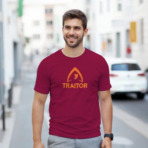 May include: A maroon t-shirt featuring an orange graphic and the word "TRAITOR" in orange. The graphic is a triangle shape with a face inside. The t-shirt is short-sleeved.