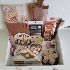 May include: A white box filled with stationery and decorative items in shades of brown and beige. Includes a notebook, stickers, pens, washi tape, a sequined heart, a flower hair clip, and coffee-themed stickers and a coffee bar sign.
