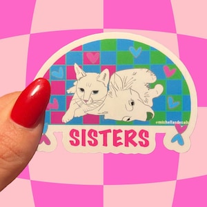May include: A sticker featuring two white cats, one upright and one upside down, with the word "SISTERS" in pink. The background is a checkered pattern of pink, blue, and green with heart accents.