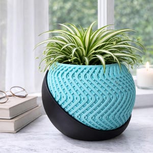 May include: A turquoise patterned plant pot with a black base, containing a green and white plant. The pot has a distinctive woven design. The plant pot is on a white surface near a window.