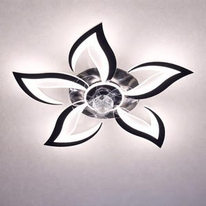 Op de afbeelding: Een moderne plafondlamp met een bloemvormig ontwerp. De lamp heeft vijf witte bloembladen met een zwarte rand, die uitstralen vanuit een centrale chroomkleurige naaf. In het midden zit een heldere, facetgeslepen kristallen lamp.