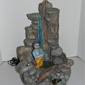 May include: A ceramic table lamp shaped like a waterfall with a fisherman figurine standing in the water. The lamp is made to look like a rocky stream with a waterfall flowing down a tree trunk. The lamp has a gold-colored base and a white cord.