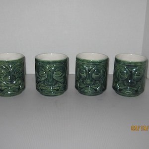 May include: Set of four green ceramic tiki mugs with carved faces. Each mug has a different facial expression.