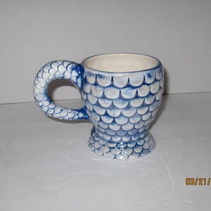 May include: A blue and white ceramic mug with a fishtail handle and a fish scale design.