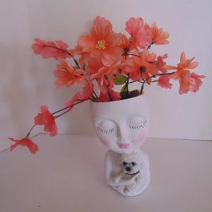 May include: White ceramic planter shaped like a woman's face with closed eyes and a small white dog in her arms. The planter is filled with pink flowers.