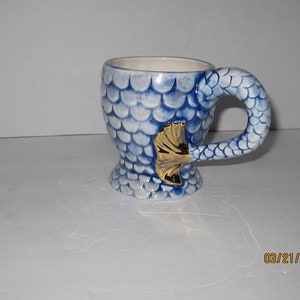 May include: A blue and white ceramic mug shaped like a mermaid tail. The mug has a gold fin and iridescent scales.