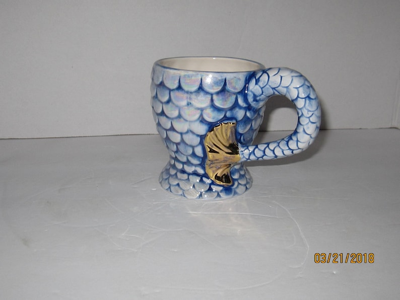 May include: A blue and white ceramic mug shaped like a mermaid tail. The mug has a gold fin and iridescent scales.