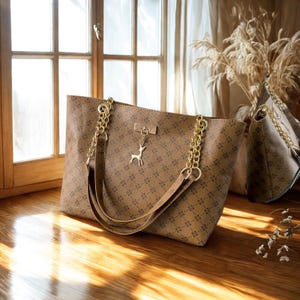 Könnte beinhalten: Eine braune Tragetasche mit geometrischem Muster und goldenen Kettenriemen. Die Tasche hat einen kleinen goldenen Hirschanhänger und einen rechteckigen goldenen Akzent. Eine zweite Tasche ist im Hintergrund zu sehen.