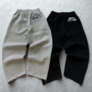 Op de afbeelding: Twee joggingbroeken, één lichtgrijs en één zwart, worden getoond. Elke broek heeft een achterzak met een cirkelvormige afbeelding. De joggingbroeken hebben een trekkoord aan de onderzoom. De lichtgrijze broek staat links, de zwarte rechts.