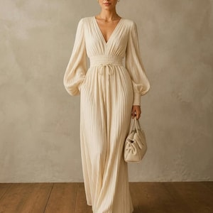 May include: A floor-length cream-coloured pleated dress featuring long bishop sleeves with cuffed wrists, a deep V-neckline, and a smocked waist with a drawstring tie. The dress is paired with a matching cream-coloured structured handbag.