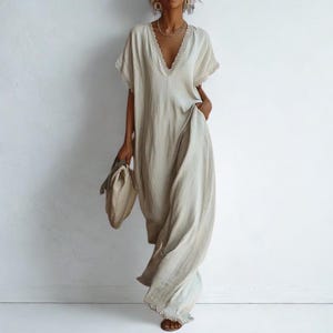 Può includere: Abito lungo beige chiaro con scollo a V e maniche corte. L'abito presenta un bordo in pizzo lungo lo scollo e le maniche. Una borsa abbinata è tenuta in una mano. L'abito ha tasche ed è realizzato in un tessuto fluido.