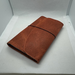 May include: A folded, brown leather journal with a black elastic band closure. The leather has a textured appearance. The journal is rectangular and appears to be closed. The background is white.