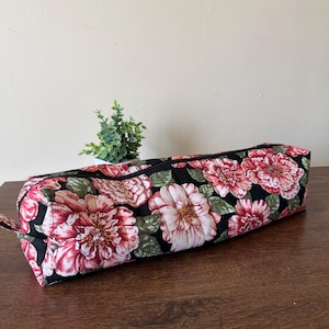 May include: A long, rectangular fabric pencil case featuring a black background with a repeating floral pattern of large, pink and white peonies and green leaves. The case has a black zipper closure running along the top and quilted stitching.