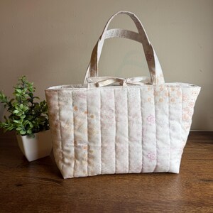 May include: A quilted tote bag featuring a soft cream background with delicate floral patterns in shades of pink and orange. The bag has vertical stitching, two fabric handles, and a small tie closure at the top center of the opening.