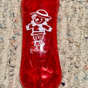 May include: A red, translucent bottle featuring a white cartoon character in a hat and collar. The bottle has a rounded shape, possibly made of plastic or glass. The design is likely a decorative element.