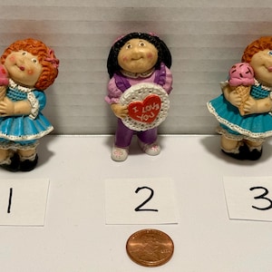 May include: Three small figurines featuring two figures with red hair in blue dresses holding pink ice cream cones and one figure with black hair in a purple outfit holding a white circular plaque with a red heart and the text I LOVE YOU.