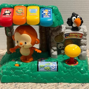 May include: A VTech Learn and Dance Interactive Zoo toy featuring a green base with a central orange monkey figure, four colourful animal buttons labelled Penguin, Parrot, Elephant, and Giraffe, a black toucan, and a yellow button on a red post.