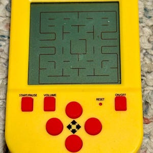 May include: A yellow Pac-Man handheld game console. The screen shows the maze game. It features red buttons for start/pause, volume, reset, and on/off, along with a directional pad. The Pac-Man logo is at the top.