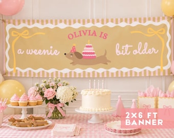May include: A rectangular party banner with a tan background, white scalloped borders, and yellow ribbon graphics. It features the pink text 'OLIVIA IS' and white text 'a weenie bit older' with a dachshund wearing a party hat and carrying a cake.